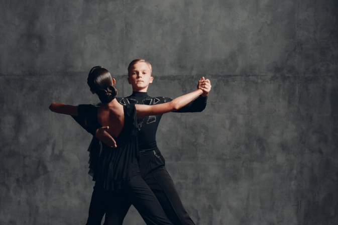 Due persone ballano con grazia in abiti formali contro un muro grigio strutturato. La ballerina a sinistra indossa un abito nero, mentre quella a destra indossa un abito nero a maniche lunghe. Stanno ballando abbracciati, eseguendo un ballo da sala.