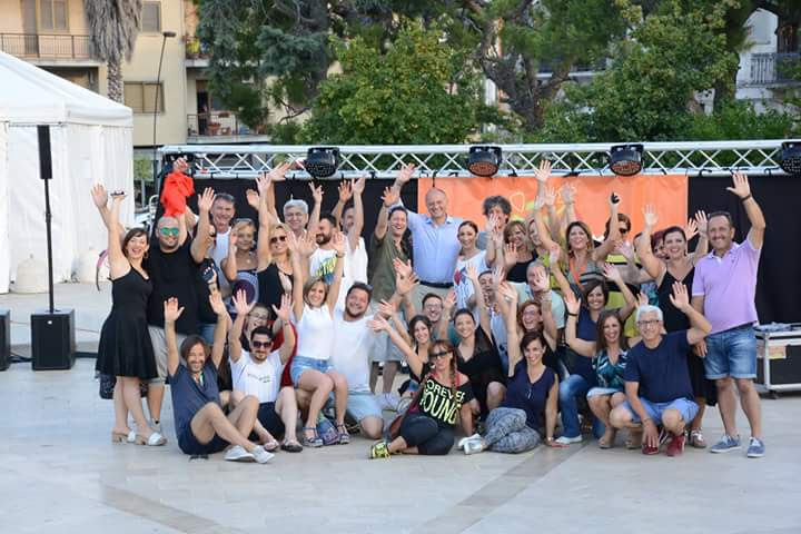 Un gruppo di quindici persone sta in una stanza chiusa su un pavimento di legno, sorride e posa per la macchina fotografica. Indossano una varietà di abiti, che vanno dal casual al semi-formale. Lo sfondo è una stanza semplice e ben illuminata.