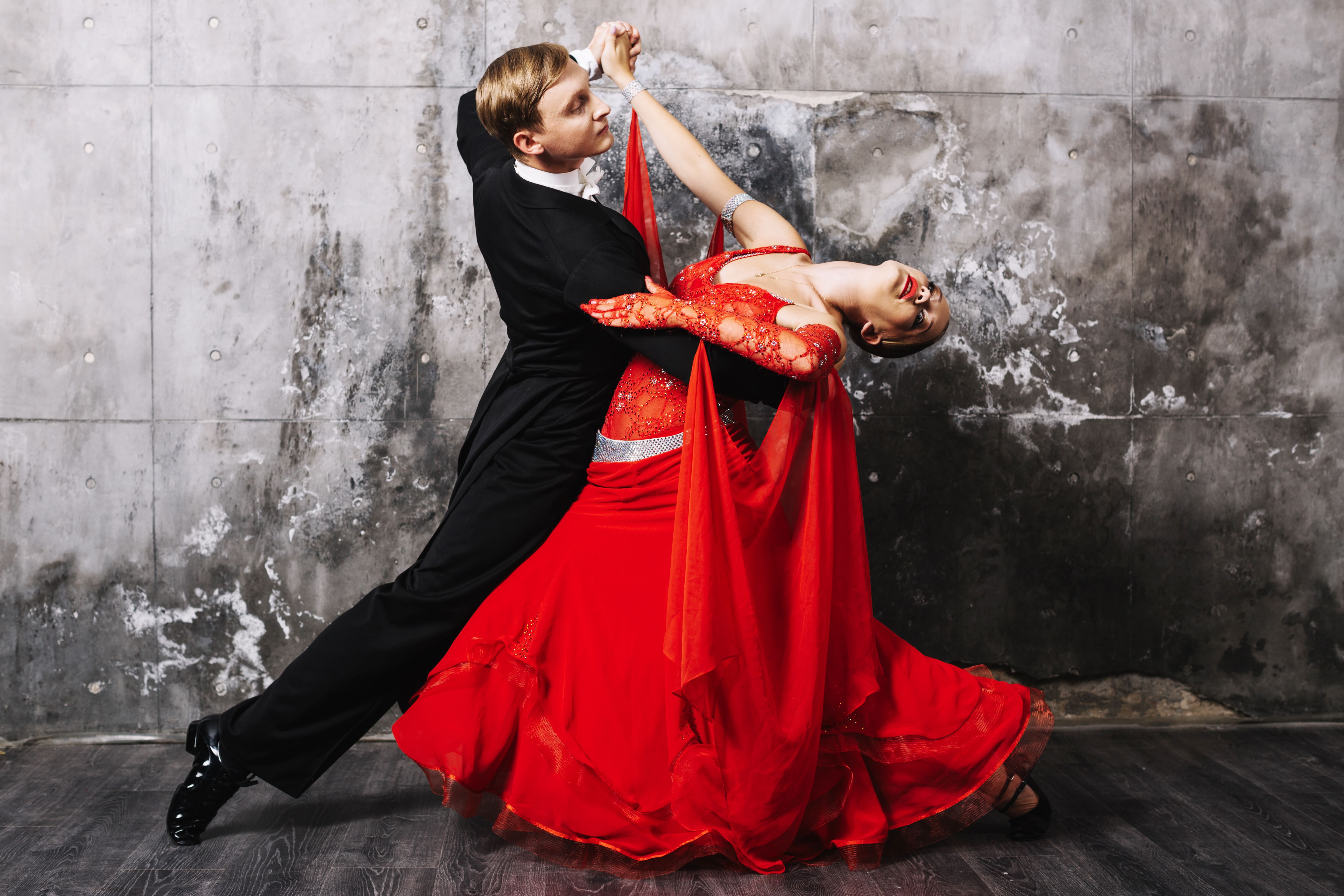 Una coppia esegue una danza elegante. La donna, in un fluido abito rosso, ha un piede sollevato mentre si avvicina con grazia al suo partner. L'uomo, in abito nero e cravatta, la tiene saldamente. Lo sfondo è scuro e sfocato, accentuando la loro posa drammatica.